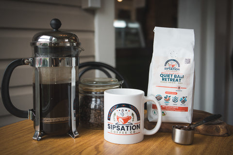 Sipsation Coffee Co. products including a French press, coffee bag, and mug on a wooden table.